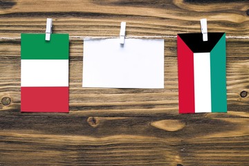 Hanging flags of Italy and Kuwait attached to rope with clothes pins with copy space on white note paper on wooden background.Diplomatic relations between countries.