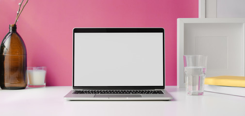 Modern workspace with open blank screen laptop computer with mock up frame and office supplies on white table and pink wall