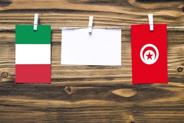 Hanging flags of Italy and Tunisia attached to rope with clothes pins with copy space on white note paper on wooden background.Diplomatic relations between countries.