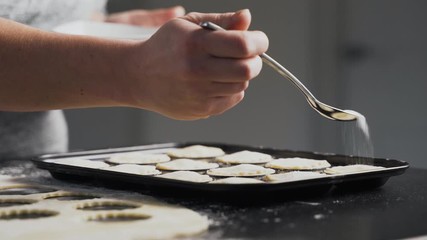 Lady sprinkles sugar on mince pies in a baking tin. Woman baking holiday dessert pastry finishes by shaking sugar from a spoon onto the dough top. Black kitchen counter with flour. ZOOM OUT.