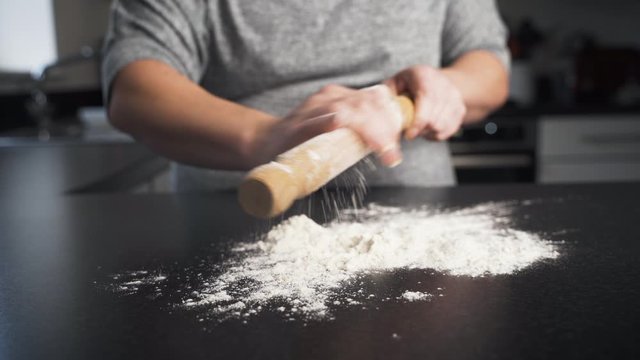 Woman prepares her rolling pin to flatten dough by wiping a coating of flour over the wooden roller. Lady baking in her kitchen gets tools ready.