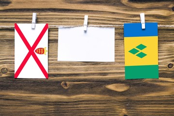 Hanging flags of Jersey and Saint Vincent And The Grenadines attached to rope with clothes pins with copy space on white note paper on wooden background.Diplomatic relations between countries.