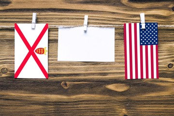 Hanging flags of Jersey and United States attached to rope with clothes pins with copy space on white note paper on wooden background.Diplomatic relations between countries.