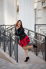 Young brunette girl weared in red dress and leather jacket with a backpack on background of the City. Fashion style and shopping walk in a city. Street style Modern strict urban woman