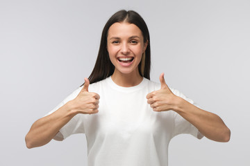 Young woman showing thumbs up with positive emotions and happiness, isolated on gray background....