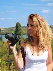 Long-haired blonde sexy woman with a SLR camera. Girl in white t-shirt and blue jeans, sitting cross-legged on a low wall in the sun.