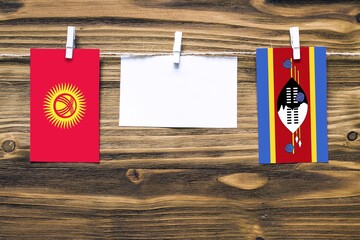 Hanging flags of Kyrgyzstan and Swaziland attached to rope with clothes pins with copy space on white note paper on wooden background.Diplomatic relations between countries.