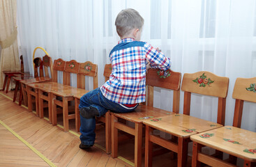 Cute alone caucasian baby boy with fair hair sits on the chair, back to the viewer, face to the window. Loneliness and children melancholy concept. Indoors, copy space.