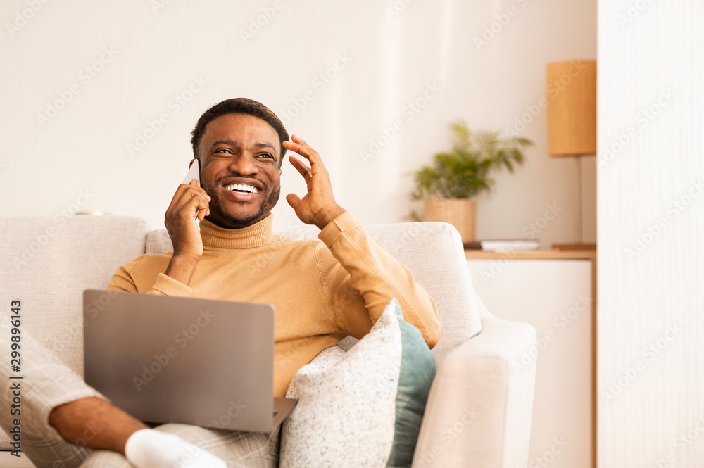 Poster man using laptop talking on phone sitting on sofa indoor