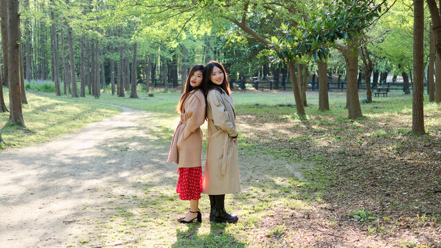 Two beautiful Chinese young girls standing back to back with hands in pocket and looking at camera in sunny autumn forest park, best friends posing outdoor.