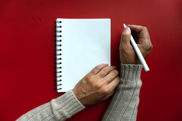 Mans right hand in winter sweater hold white pen and writes his plans and list of motivation for future for new year 2020 in white papered blank spiral notebook, red backfround