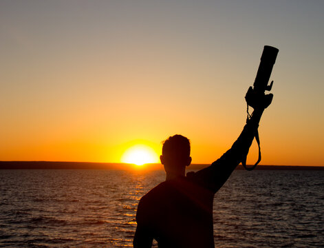 Silhouette photograph of the man is standing back on the shore of the sea and holds a camera in his raised hand with fully happy moment with beautiful sunset light.