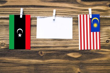 Hanging flags of Libya and Malaysia attached to rope with clothes pins with copy space on white note paper on wooden background.Diplomatic relations between countries.