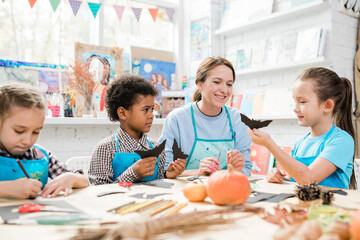 Diligent schoolkids making halloween toys and showing them to happy teacher