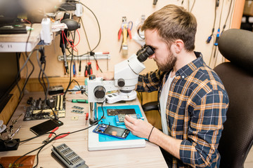 Bearded repairman sitting by table in front of microscope to see tiny details