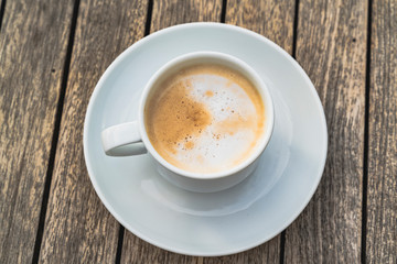 Hot foaming cappuccino in a white ceramic cup on a saucer stands on a wooden table, top view