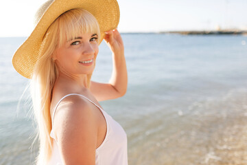 Beautiful blonde woman in white dress and straw hat on the beach