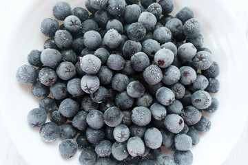  White plate with frozen chokeberry berries on a wooden white table. Close-up. 