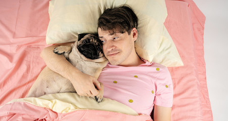 Handsome man lying with pug at bed. Young man in a bed under a rug with his dog in an embrace....