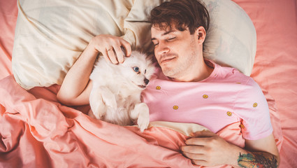 Handsome man lying with little white dog with blue eyes at bed. Young man in a bed under a rug with his dog in an embrace. Handsome young hipster with his dog resting on bed at home