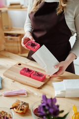 Hands of woman putting handmade pink soap bar into white paper packet