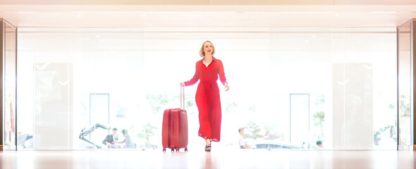Full-length portrait of gorgeous lady with blonde hair wearing red jumpsuit entering in the bright hotel lobby with a suitcase. Business trip concept. Horizontal shot. Front view