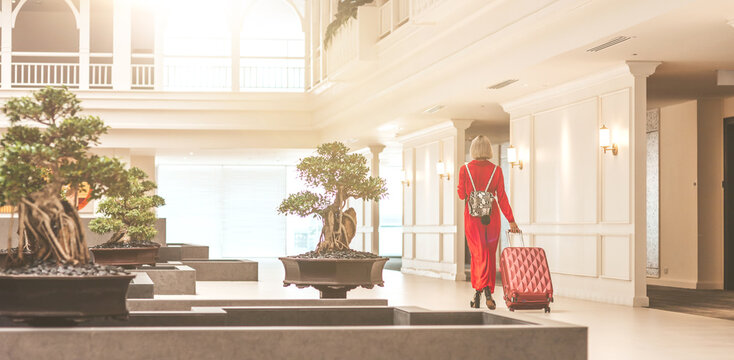 Full-length portrait of gorgeous lady with blonde hair wearing red jumpsuit entering in the hotel lobby with a backpack and a suitcase. Business trip concept. Horizontal shot. Back view