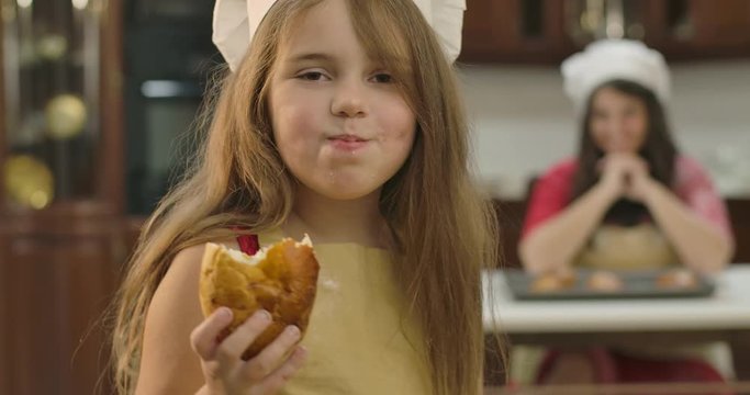 Small Caucasian daughetr looking back at her mom,. turning to camera and chewing sweet baked cookie. Little cute girl eating bun at the kitchen at home. Cinema 4k footage ProRes HQ.