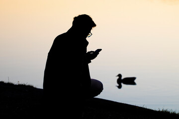 Young man checking mobile phone near lake with duck