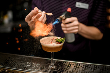 Bartender adding to an alcoholic cocktail in the glass spices from the spoon and burning them