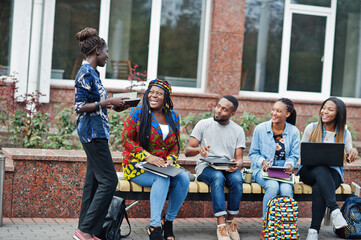 Group of five african college students spending time together on campus at university yard. Black afro friends studying at bench with school items, laptops notebooks.