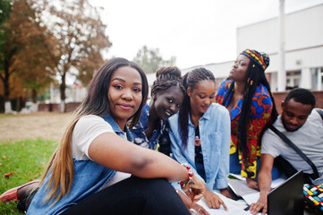 Group of five african college students spending time together on campus at university yard. Black afro friends sitting on grass and studying with laptops. - obrazy, fototapety, plakaty