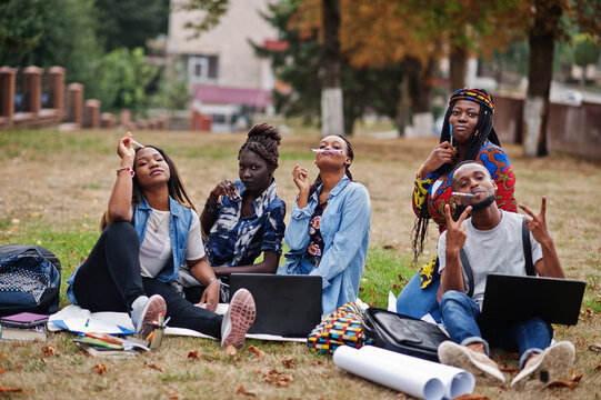 Group of five african college students spending time together on campus at university yard. Black afro friends sitting on grass and studying with laptops.