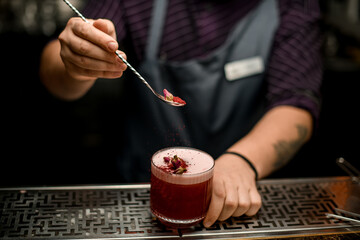 Male bartender decorating a delicious alcoholic cocktail in the glass with a dried rose buds on the spoon