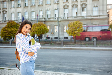 Photo of college student girl, smiling at camera, pressing book to chest, wearing backpack, ready to go to studies, start new project and suggest new ideas.