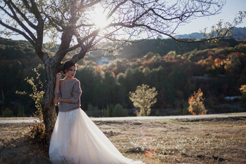 Young beautiful lonely bride posing on the nature and look to the lake and forest. Female wearing wedding dress and sweater at the background trees against the sunset.