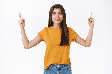 Pleased, good-looking tender young woman in yellow t-shirt pointing fingers up, smiling with satisfied amused expression, checking out interesting proposal or promo, white background