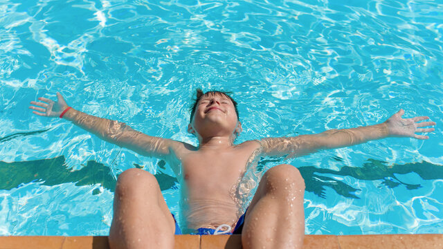  Portrait of cheerful European boy swimming in pool. He is enjoying his summer vacations. He is laying on his back with wide open hands on the water surface and smiling.