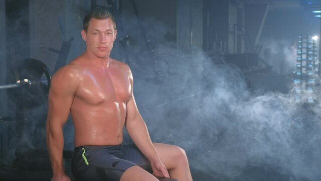 Portrait of professional muscular athletic man bodybuilder in shorts is sitting on bench in gym and looking at camera. Perfect body with naked torso. White smoke in gym in dark. Sport motivation.