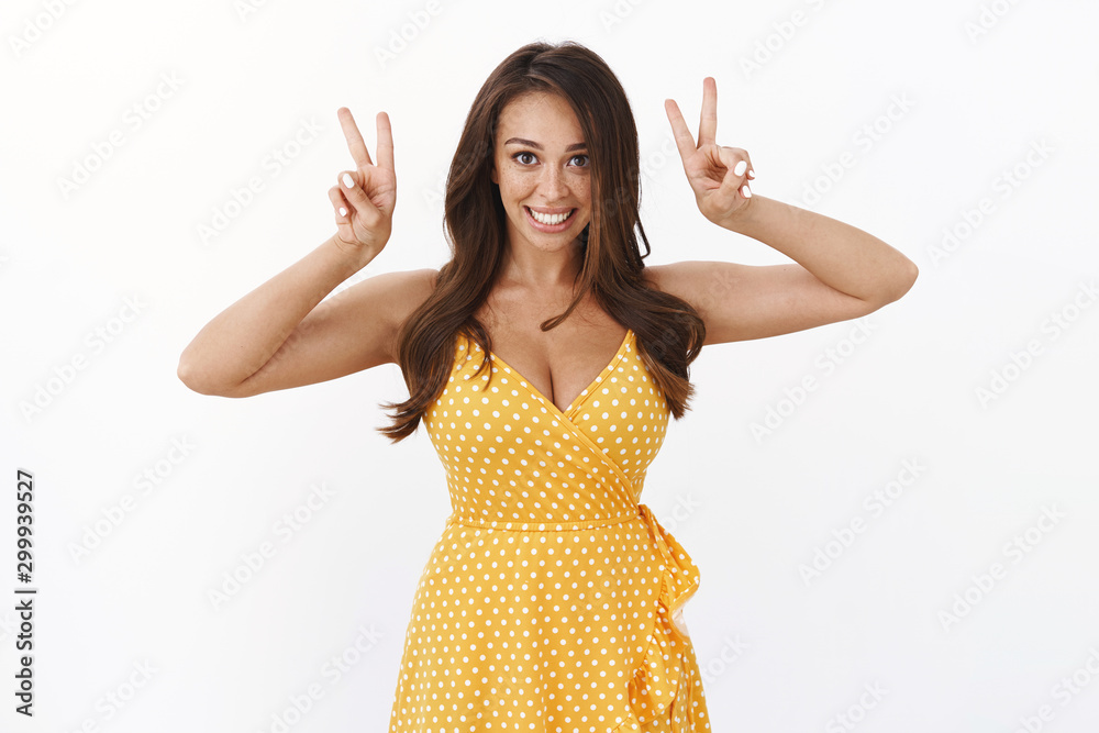 Wall mural Cheerful enthusiastic attractive girlfriend posing for cute photo from summer vacation, seaside trip, standing beautiful yellow dress show peace or victory signs, smiling delighted, white background