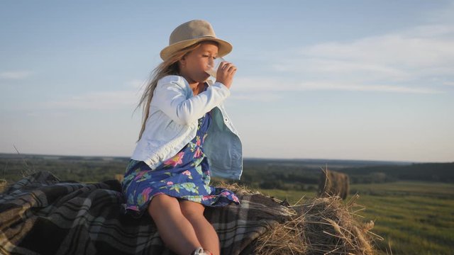 Pretty girl in blue dress drinking milk on a haystack in the field. Child on a summer picnic.