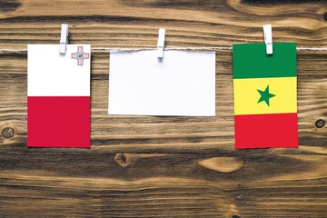 Hanging flags of Malta and Senegal attached to rope with clothes pins with copy space on white note paper on wooden background.Diplomatic relations between countries.