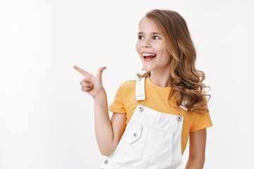 Cheerful amused cute little girl with blond hair in summer dungarees and yellow t-shirt, having fun feel entertained observe awesome interesting museum, turn left pointing copy space, smiling