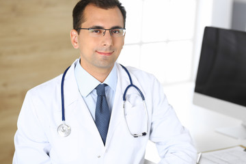 Doctor man sitting at the desk at his working place and smiling at camera. Perfect medical service in clinic. Happy future of medicine and healthcare