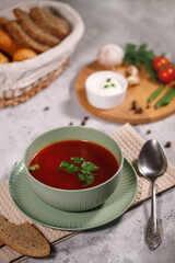 Tasty and hearty dinner. A plate with borsch on the table, next to the board are vegetables, parsley, dill, green onions, garlic, chili peppers, cherry tomatoes, and bread.