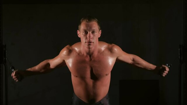 Man bodybuilder training chest muscles exercise using cable crossover machine and looking at camera, front view. White smoke in gym in dark. Sport motivation. Fit model with perfect body, naked torso.