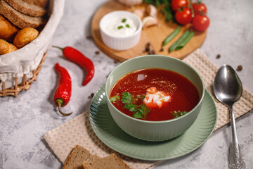 Tasty and hearty dinner. A plate with borsch on the table, next to the board are vegetables, parsley, dill, green onions, garlic, chili peppers, cherry tomatoes, and bread.