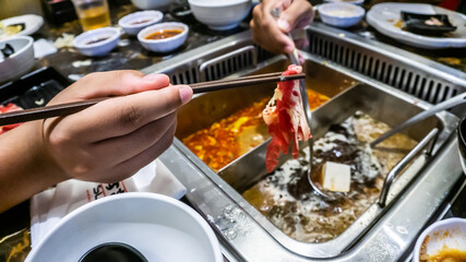 The close up view of chopstick holding a slice of raw meat before dip into the boiling soup.