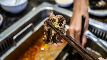 The close up view of chopstick holding a slice of cooked meat after dip into the boiling soup.