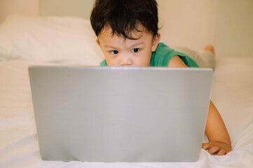 A male toddler is playing with a laptop while on the bed